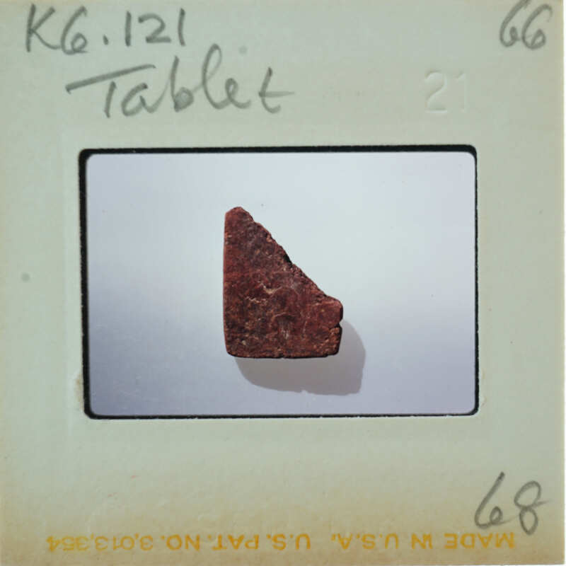 A small, irregularly shaped red fragment photographed on a white surface with a label and numbering indicating it's part of a collection or study.