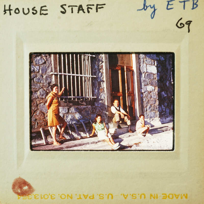 A vintage photograph of a group of people sitting outside a building with barred windows.