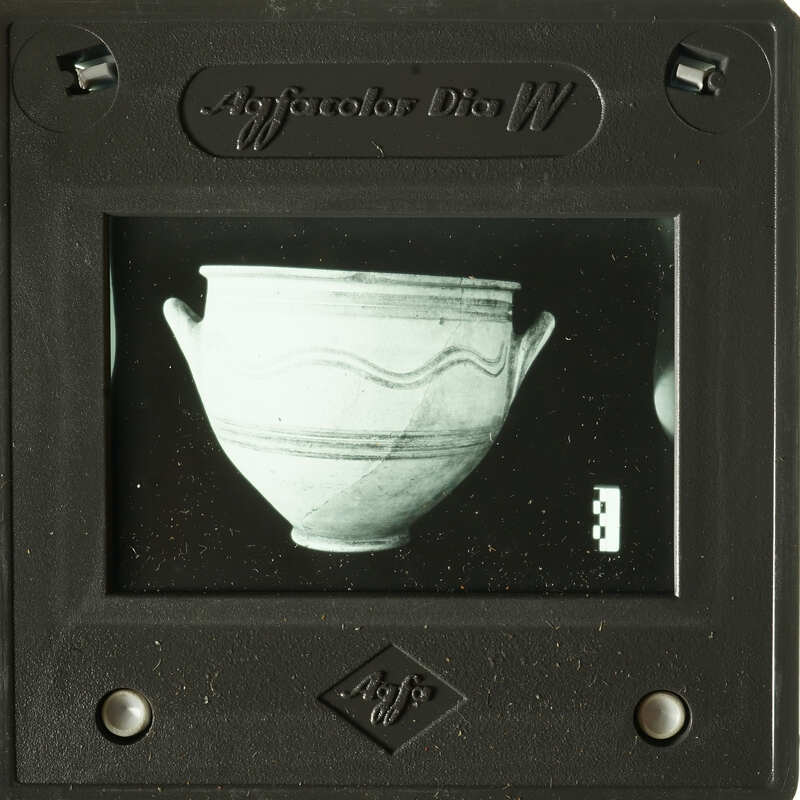 A black-and-white photograph of an ancient Greek vase displayed on a small, dark mount with visible edges and corners.