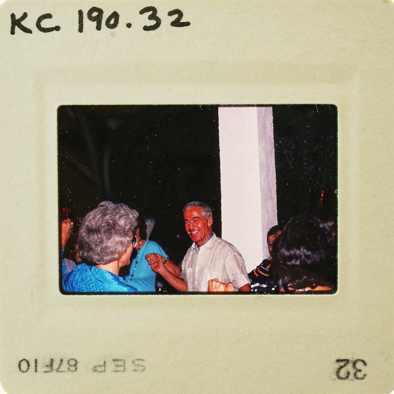 A vintage photograph of a man smiling and standing among a group of people, with the photo's border showing it is from a film roll. Catalog number KC.190.32