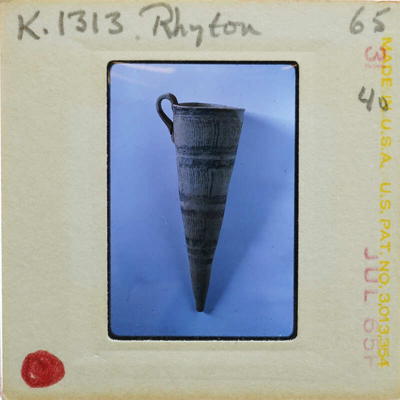 The image shows a cylindrical rhyton vase (K.1313) with a handle, displayed against a blue background.