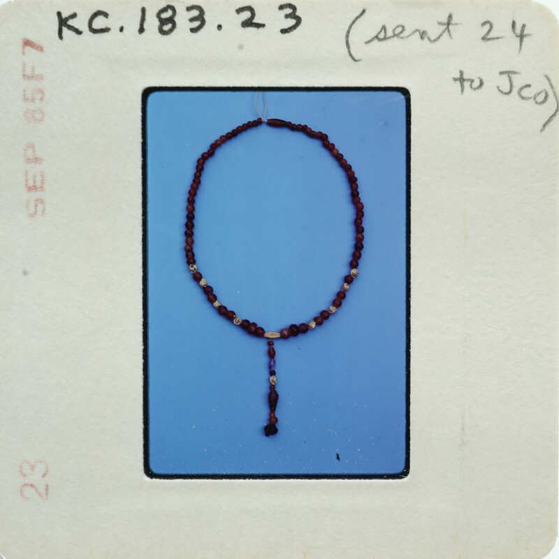 The image shows a bead necklace with a pendant against a blue background, with a white label containing handwritten notes and a barcode.
