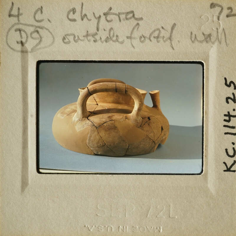 Ancient clay pot with handle and spout, displayed on light gray surface.