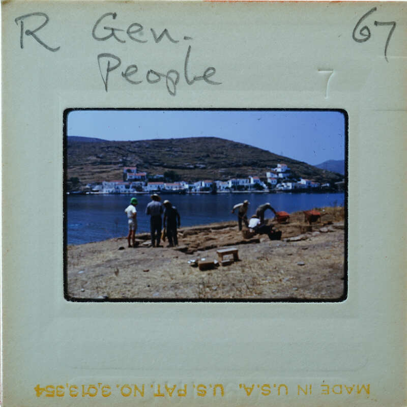 A vintage photo of people standing on a rocky shore near the water, with mountains and houses in the background.