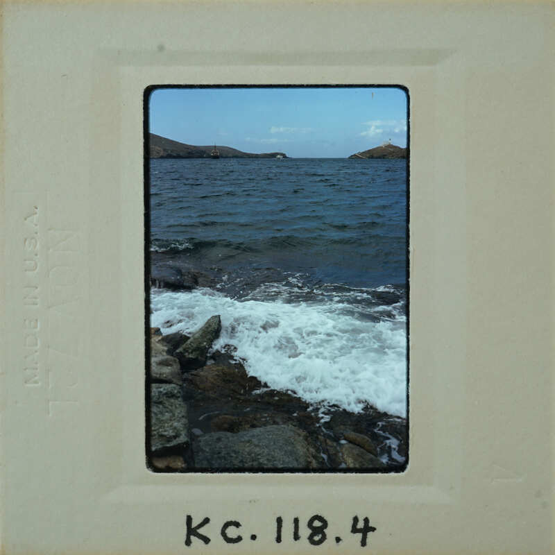 A vintage photo of a rocky shoreline with waves and an island in the background.