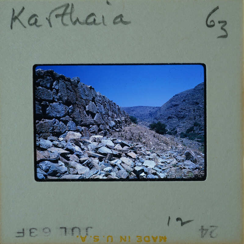 Old photograph of a stone wall with mountains and blue sky in the background, labeled "Karthaia".