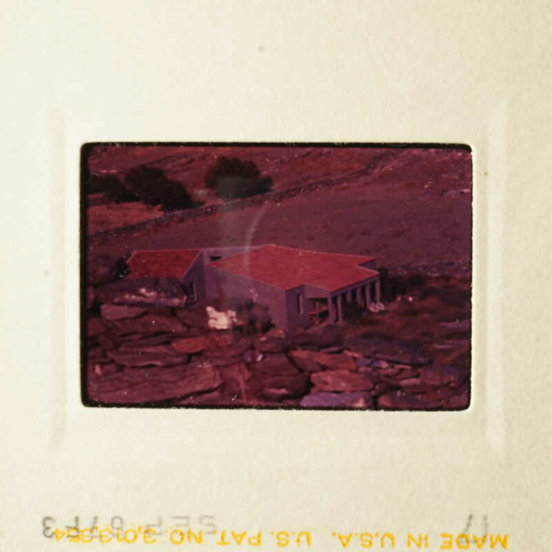 A vintage photo of a house with a red roof, set against a backdrop of hills and trees.