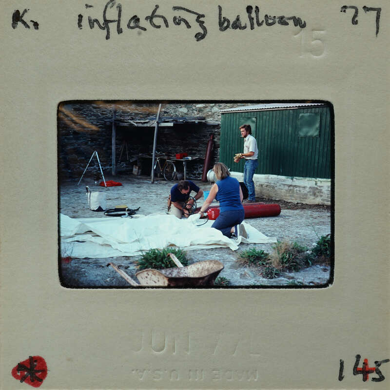 A vintage photograph of three people working on an inflatable object, with a handwritten note and date on the bottom.