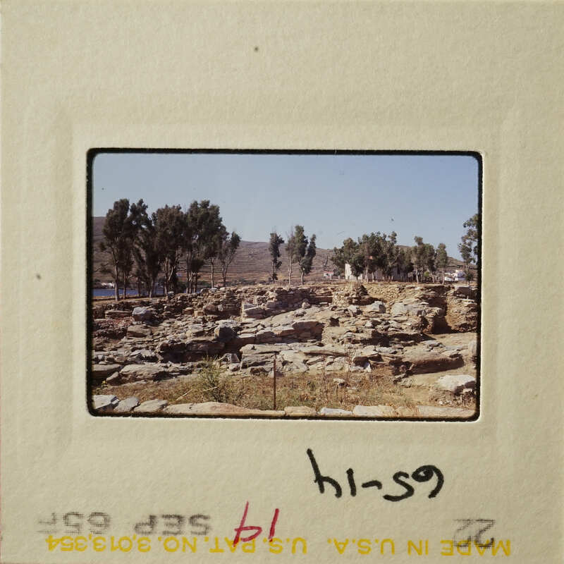 The image shows an archaeological site with ruins and trees under a clear sky.