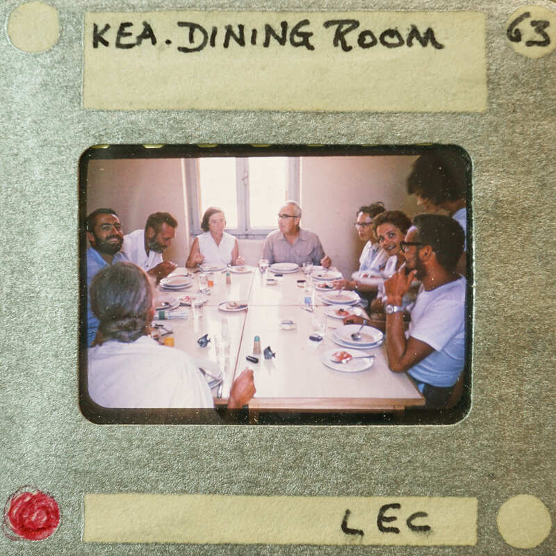A vintage photograph of a group of people gathered around a dining table, with some looking at the camera and others engaged in conversation.
