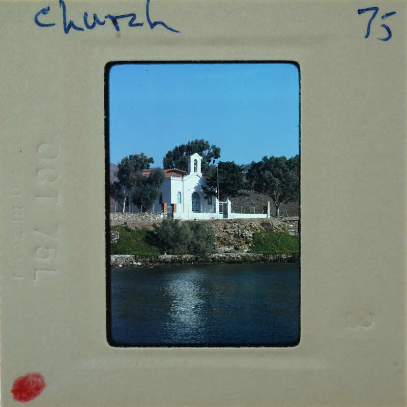 A vintage photo of a church near water, with trees and mountains in the background.