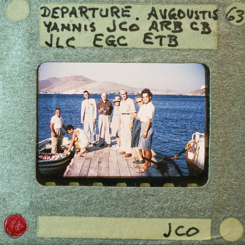 Old photograph of a group of people on a dock by the sea with mountains in the background.