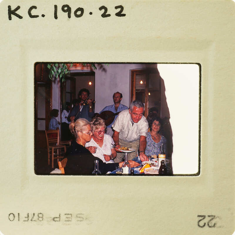 Old photo of a group of people sitting around a table, with some standing and smiling at the camera. Catalog number KC.190.22