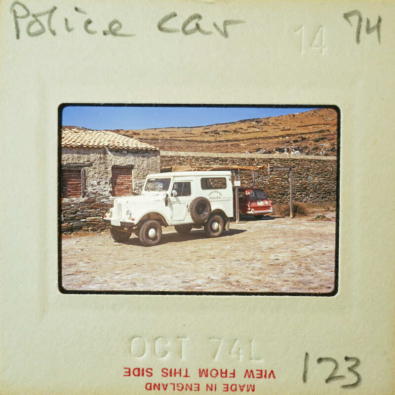 A vintage photo of a police car parked next to a stone building with mountains in the background.