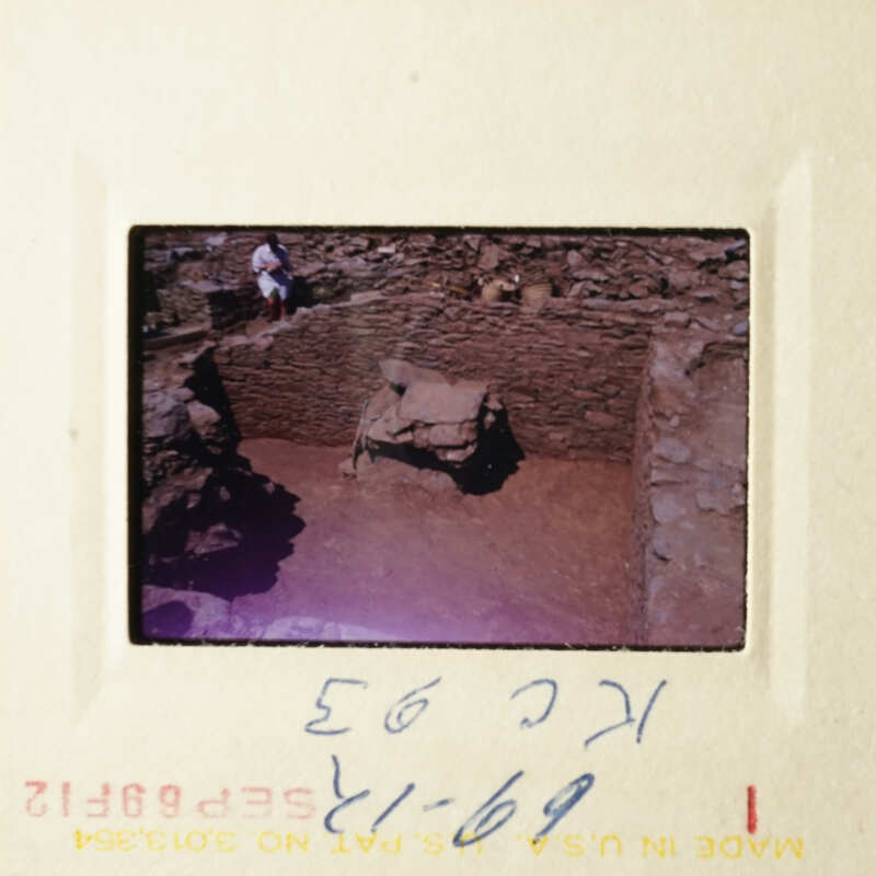 A photograph of an archaeological site with stone ruins, taken from a high angle and showing a person standing on a mound of earth.