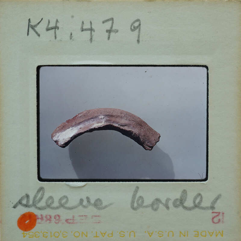 This is an image of a fossilized bone fragment with a label and handwritten notes on it, suggesting it's from an archaeological or paleontological collection.