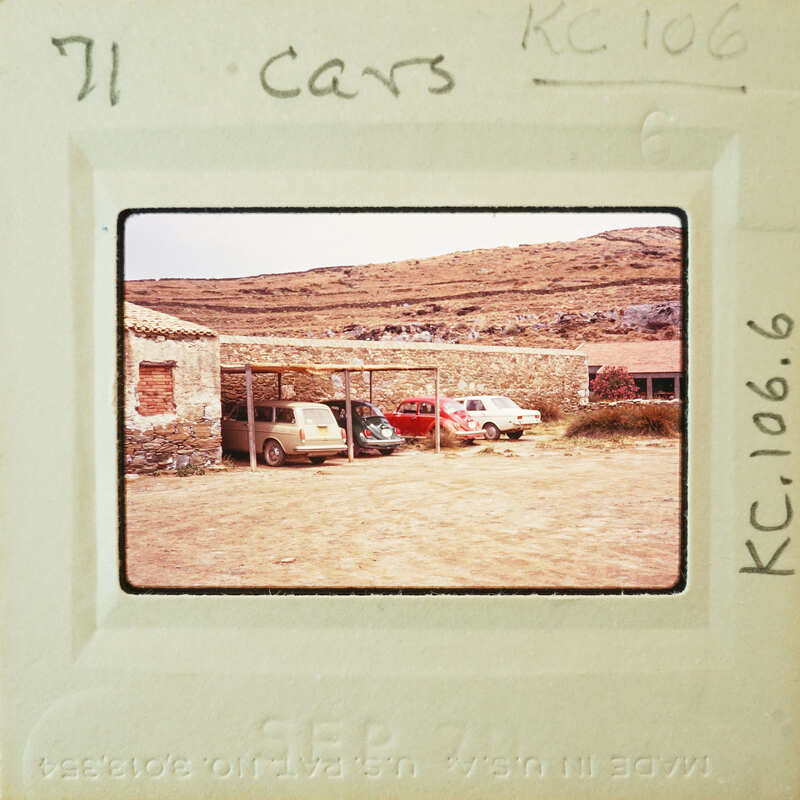 Old cars parked outside a stone building with mountains in the background.