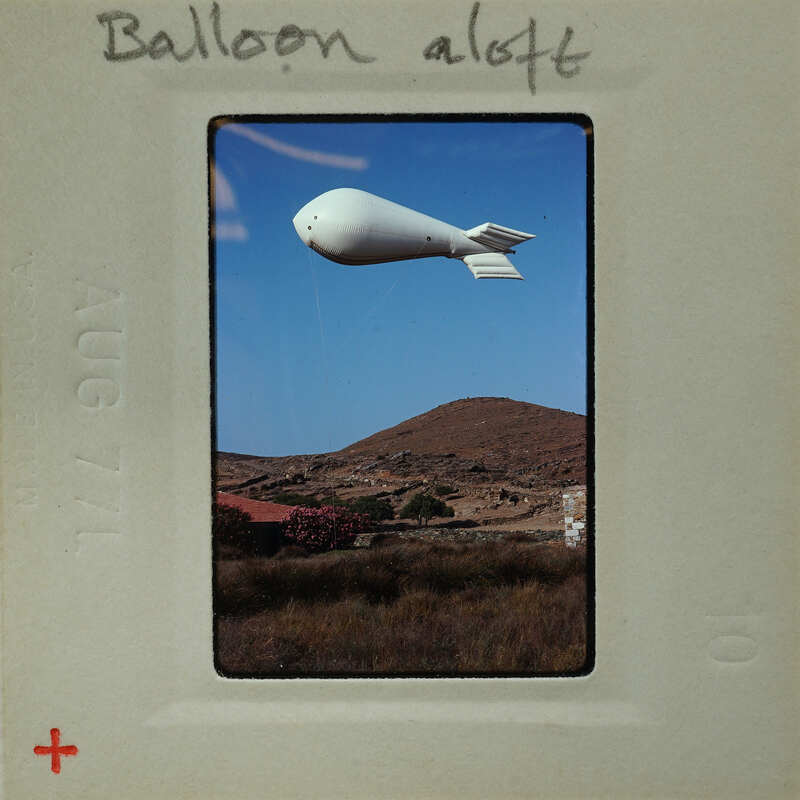 Photo of a large white blimp flying over a mountainous landscape.