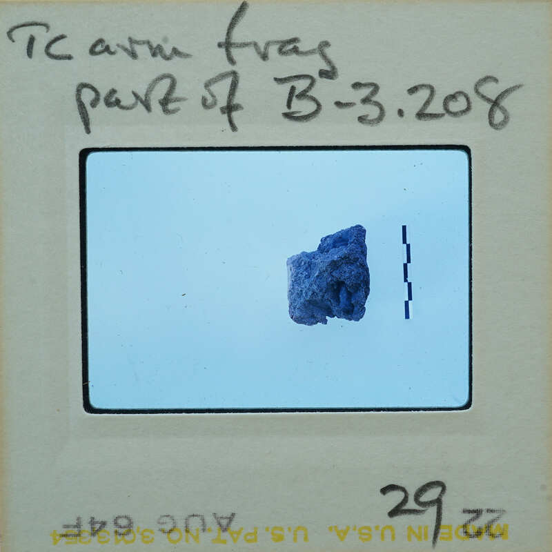 A small, dark rock sample on a light background with a label and handwritten notes.