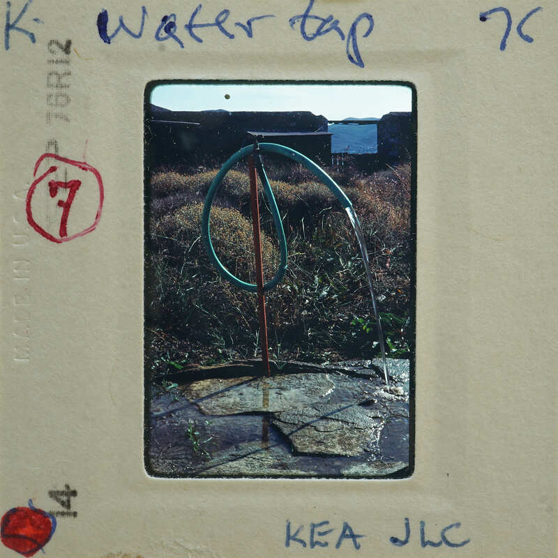 A photograph of a garden hose coiled on a metal stand, with water dripping from it onto stone tiles.