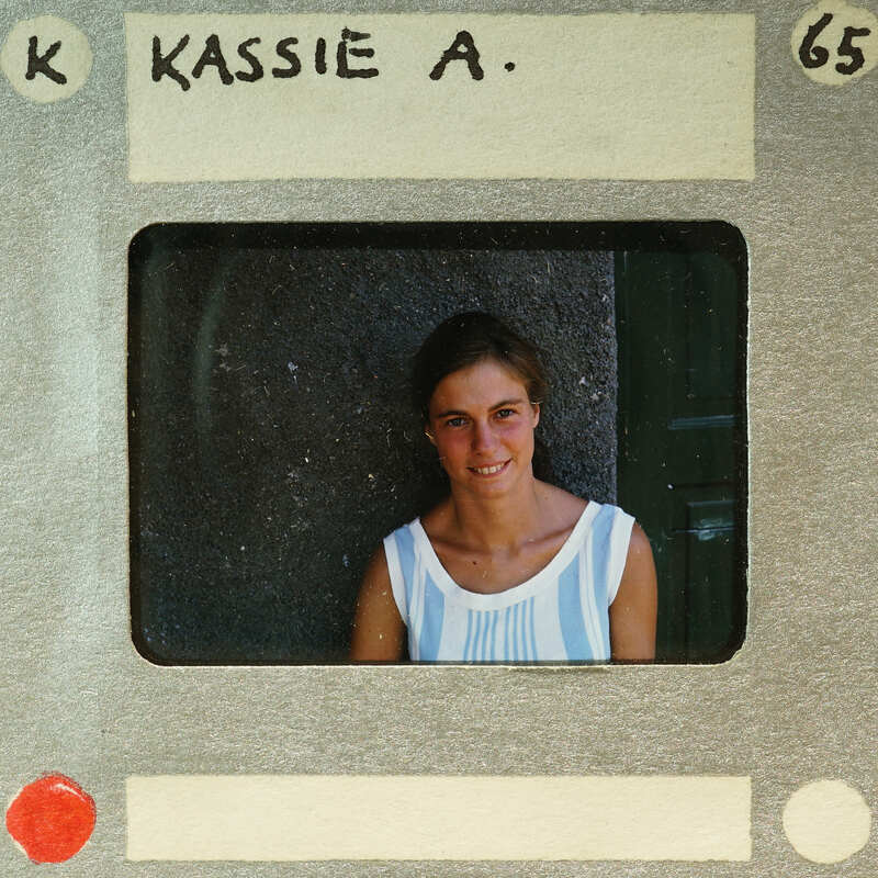 A young woman with a smile on her face, wearing a striped top and standing against a wall.