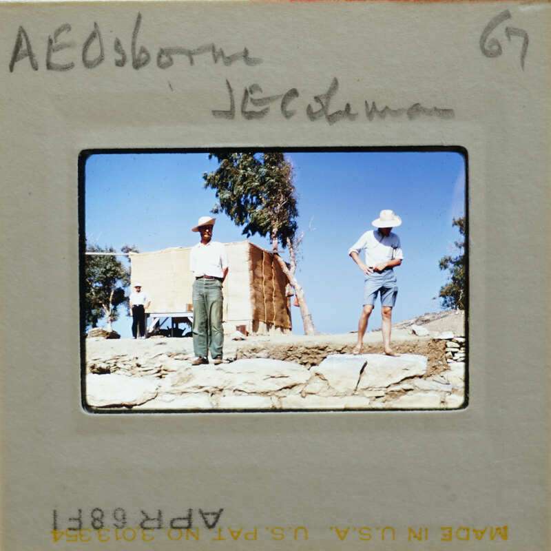 Two men standing on a rocky hillside with trees and a clear sky.