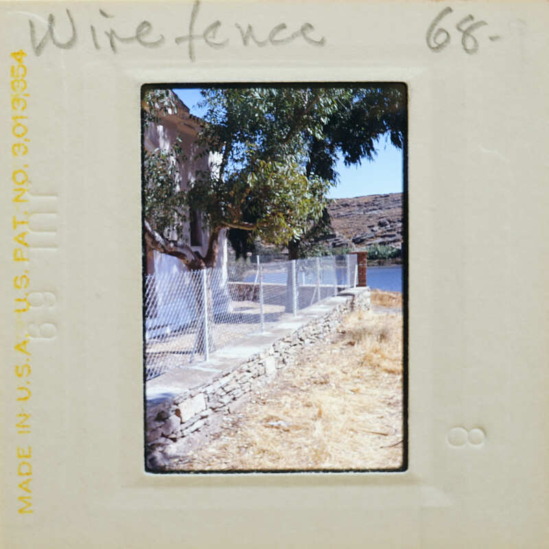 A photograph of a wire fence with trees and mountains in the background, taken during daylight.