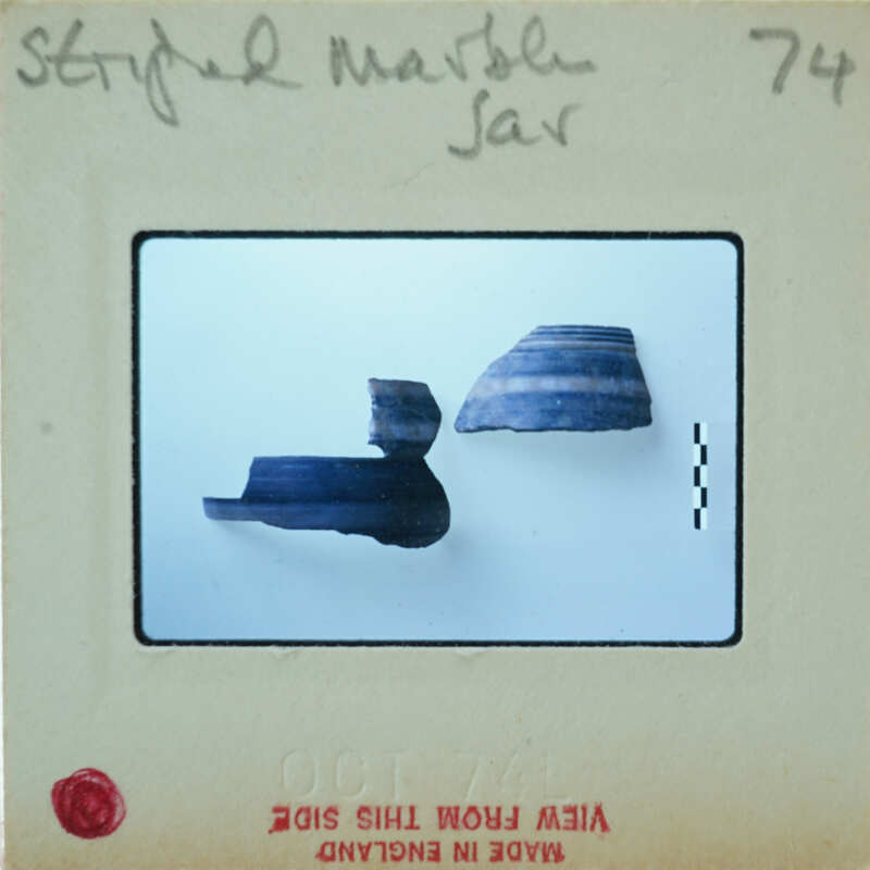 The image shows a photograph of an ancient artifact, specifically a piece of pottery with a handle and base, displayed on a white background. The photo is taken from a slightly elevated angle to show the object's profile.