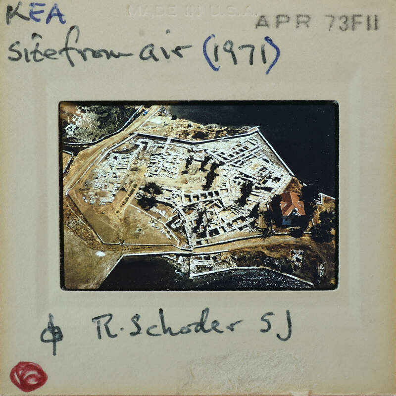 The image is a vintage photograph of an island with buildings and vegetation, taken from above.