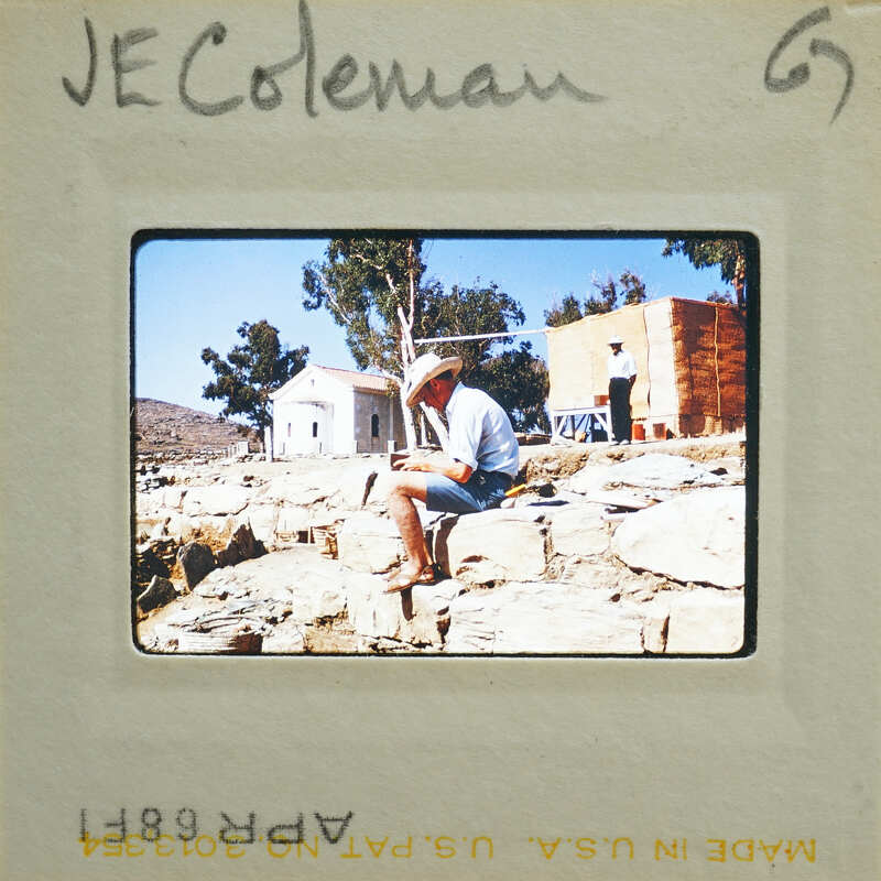 Man sitting on rock wall, wearing straw hat and shorts, with trees and building behind him.