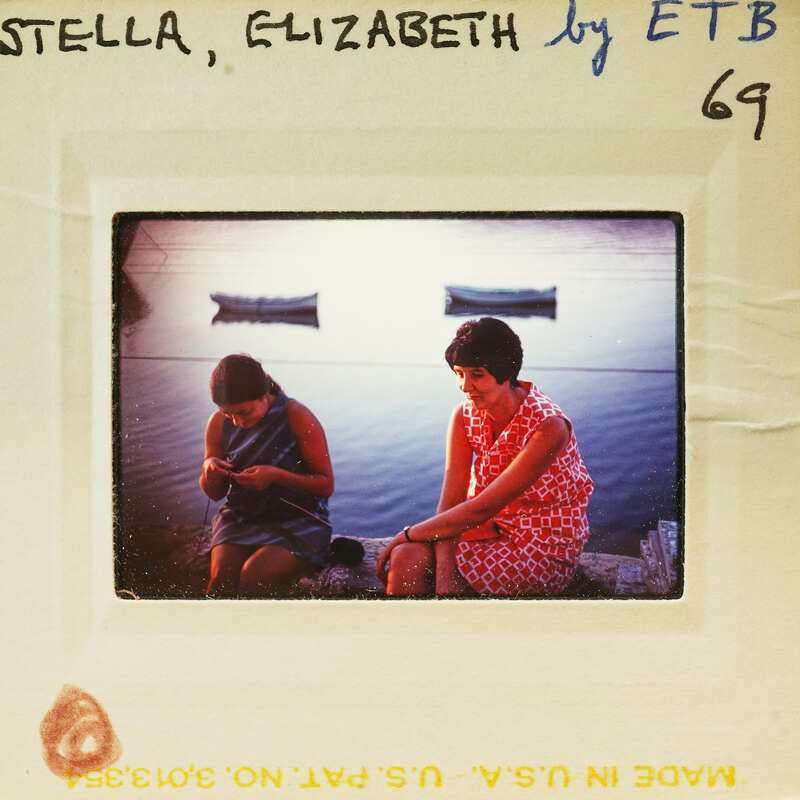 Two women sitting on a dock with boats in the background.