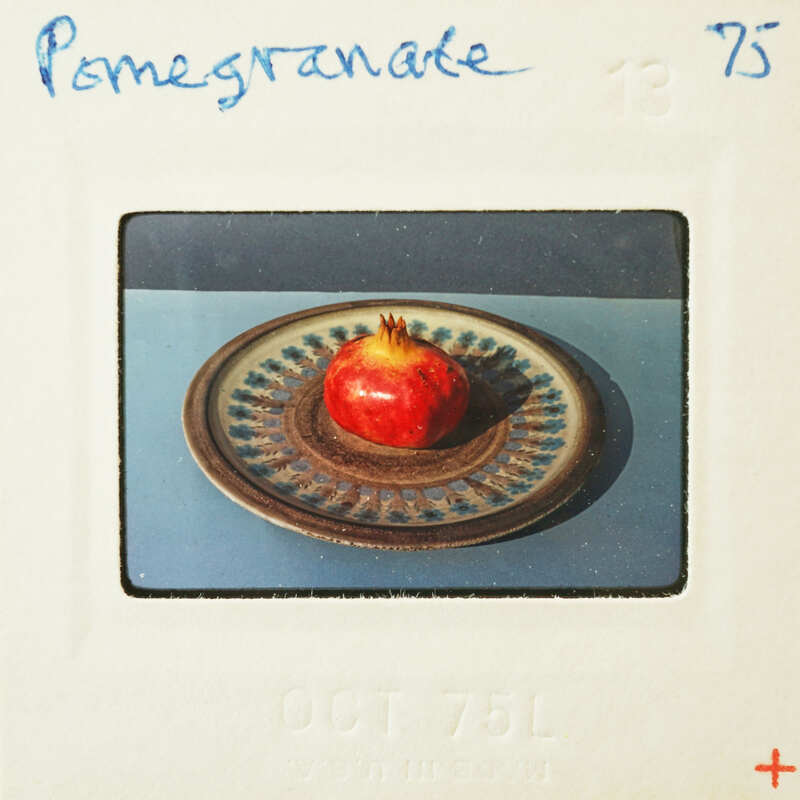 A photograph of a pomegranate on a decorative plate, enclosed within an album page with handwritten text and numbers.