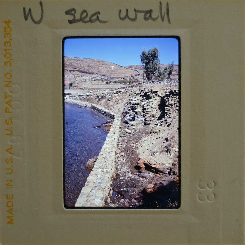 A vintage photograph of a stone wall along a river with mountains in the background.