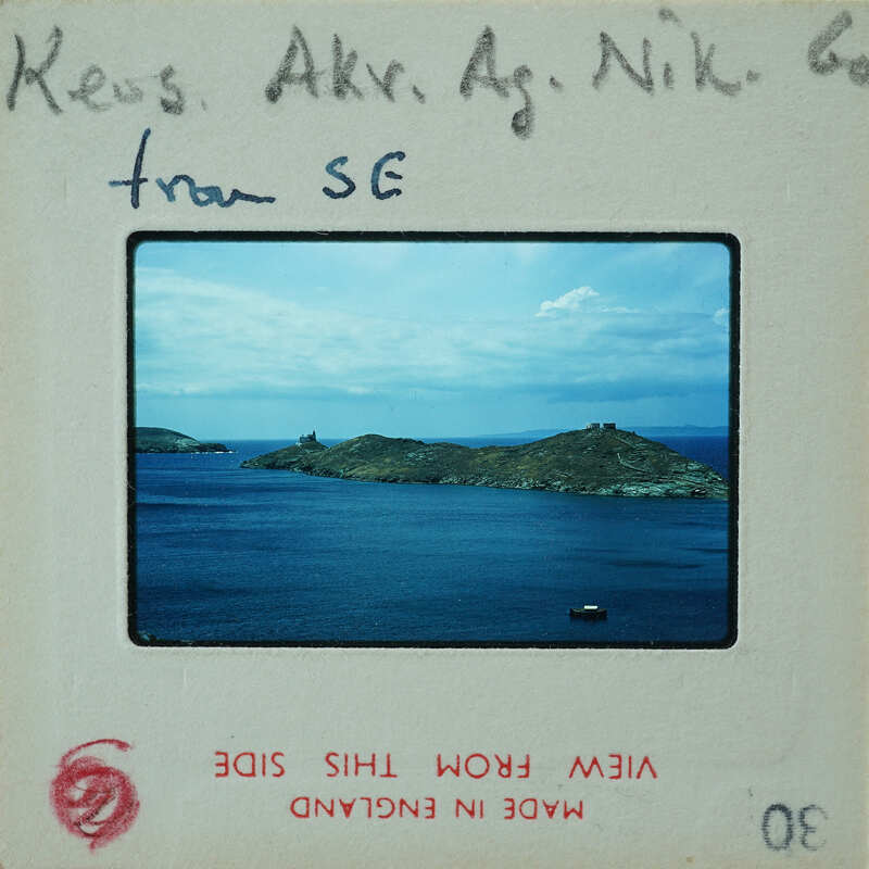 The image shows a vintage film strip with a photograph of an island and sea, accompanied by handwritten notes indicating the name "Nikos" and the location as "SE". The film strip is part of a collection or exhibit, as suggested