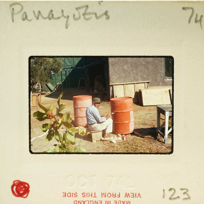 Man kneeling down next to two large orange containers, possibly barrels or tanks, with a building and trees in the background.