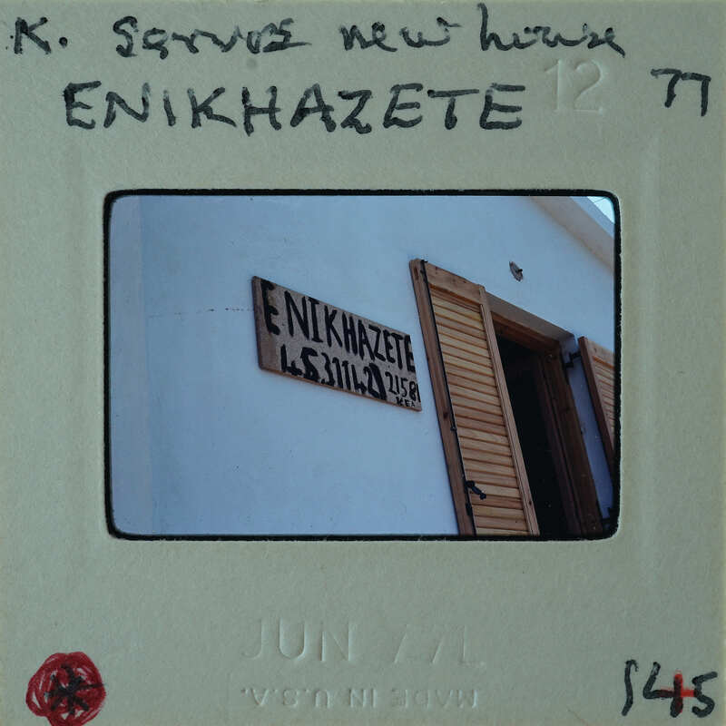 The image shows a photograph of a building with a sign that reads "ENNIHZAKEITE", which is placed on a film strip labeled with the date "JUNE 12 77" and has handwritten notes on it