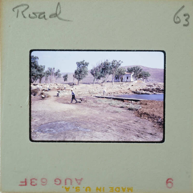 Man walking on dirt road near water with trees and mountains in the background.