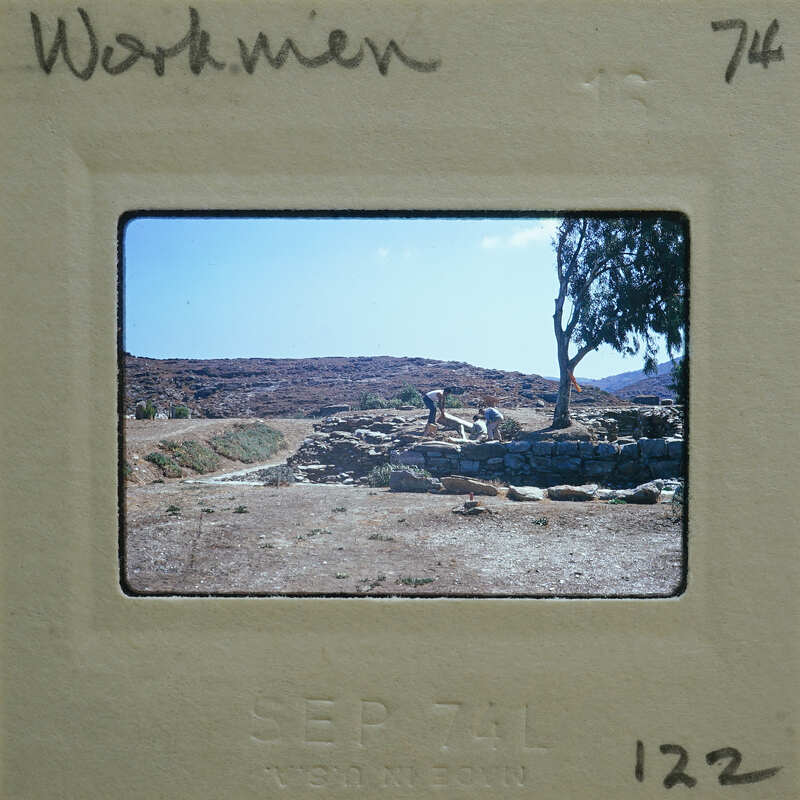 An old photo of a person working on an archaeological site with ruins and mountains in the background.