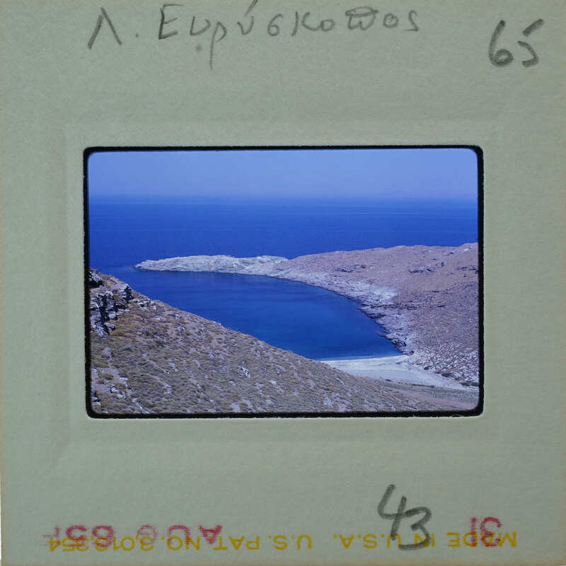 A photograph of a scenic coastal landscape with blue water and rocky terrain, possibly from an old film negative.