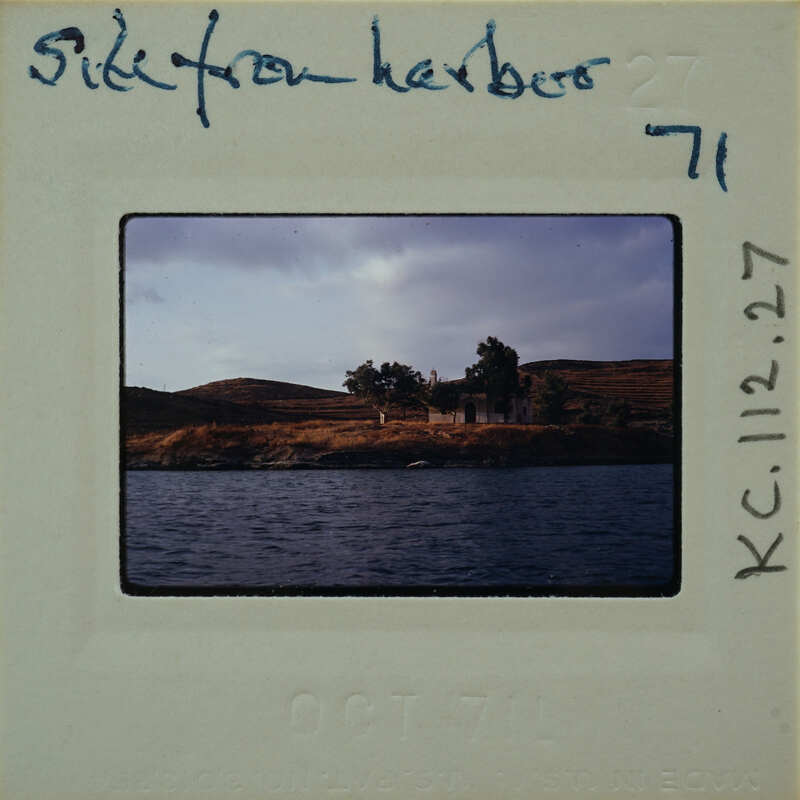 A vintage photograph of a serene landscape with water, hills, and trees, featuring an old building on the shore.