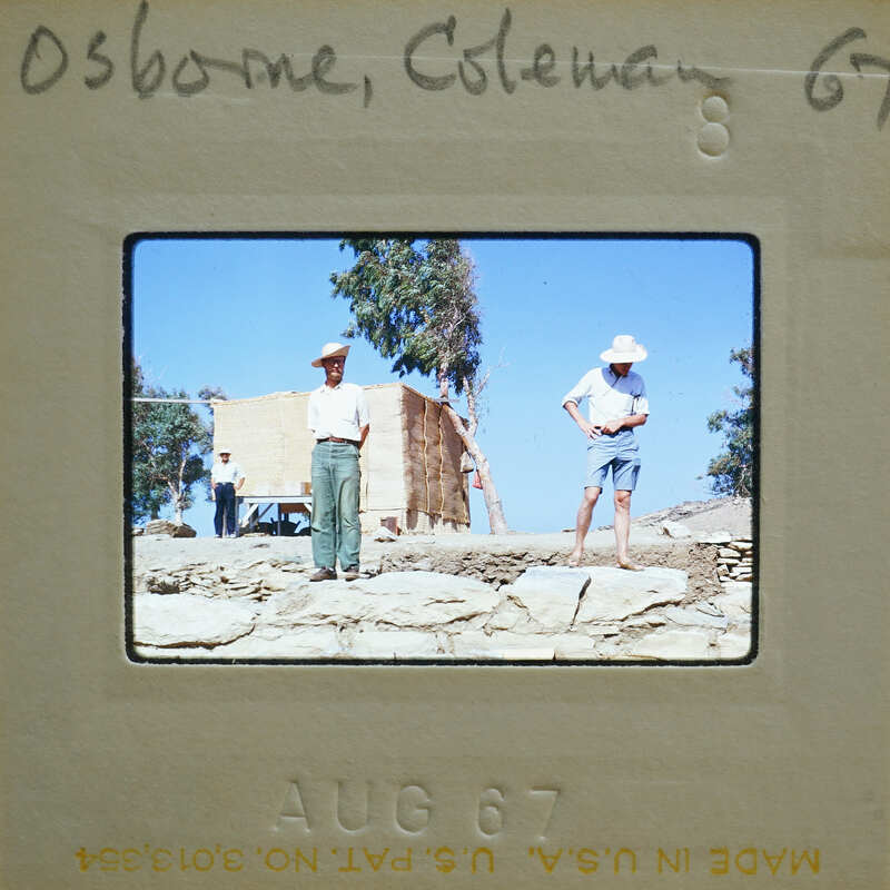 Two men standing on a rocky surface with trees and a building behind them, captured in an old photograph.