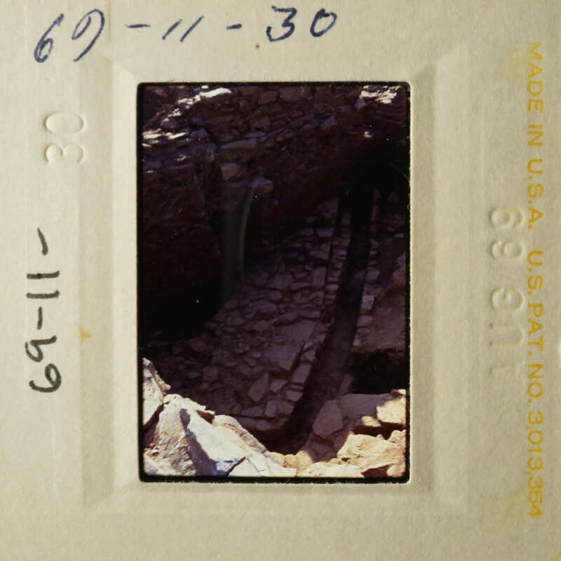 A photograph of an ancient cave entrance with stone walls and a small stream, taken from a low angle.