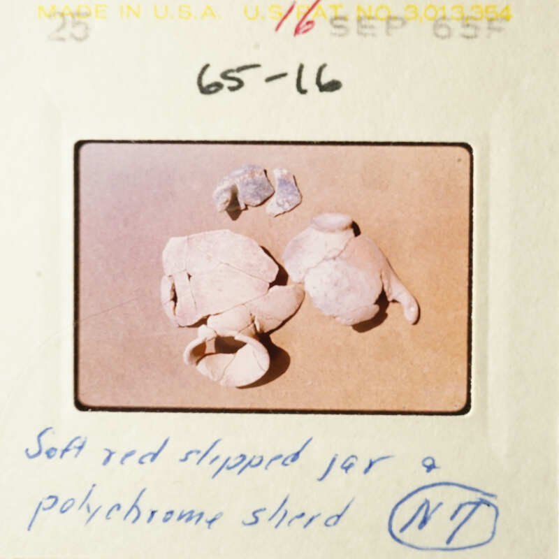 The image shows a collection of broken pottery shards on a table, with a focus on a red-rimmed shard and a small piece of red clay.