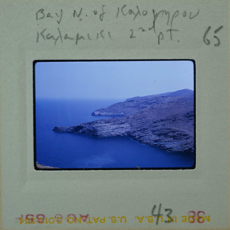 A photograph of a rocky coastline with the ocean and sky visible, on a film strip labeled "Bay N. of Ios".