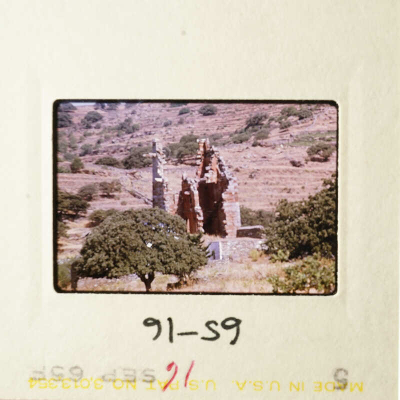 A photograph of an ancient stone structure, possibly ruins, set against a backdrop of a natural landscape with trees.