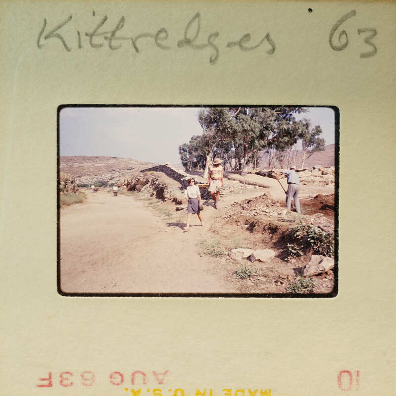 A vintage photograph of a group of people walking on a dirt road with trees and rocky terrain in the background.