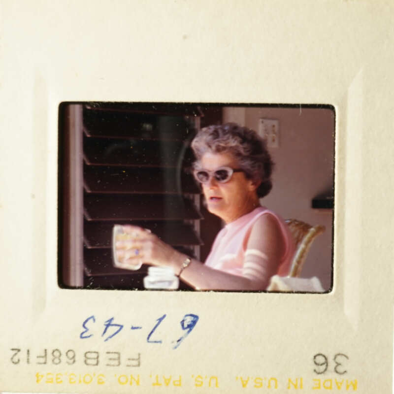 A vintage photo of an older woman holding a cup, sitting on a porch with her sunglasses on.