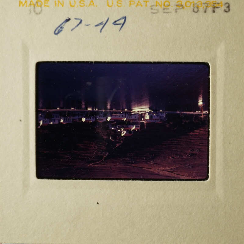A vintage photo of a nighttime scene with a boat dock and buildings, taken from a distance.