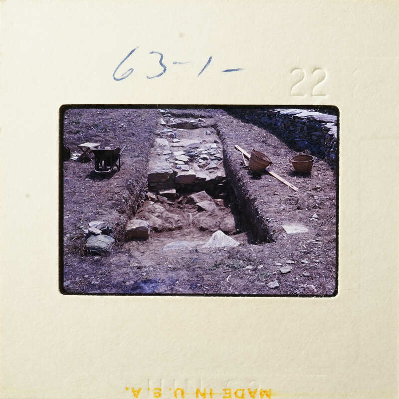 Old photograph showing an archaeological site with excavated trenches, stone foundations, and various tools.
