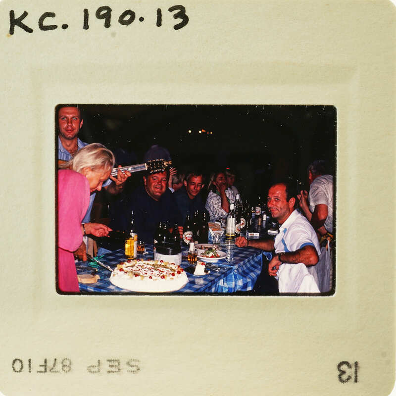 A vintage photo of a group of people gathered around a dining table with food and drinks, including a cake being cut by an older woman. Catalog number KC.190.13